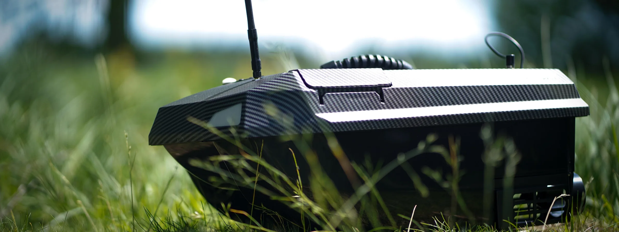 Manuowell Assassin Futterboot in Carbon-Optik im Gras am Gewässer